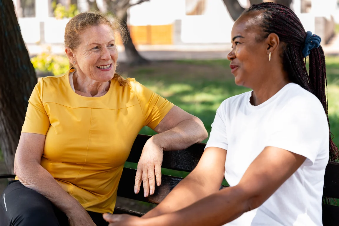 Two women of different generations and backgrounds smiling and talking warmly on a park bench