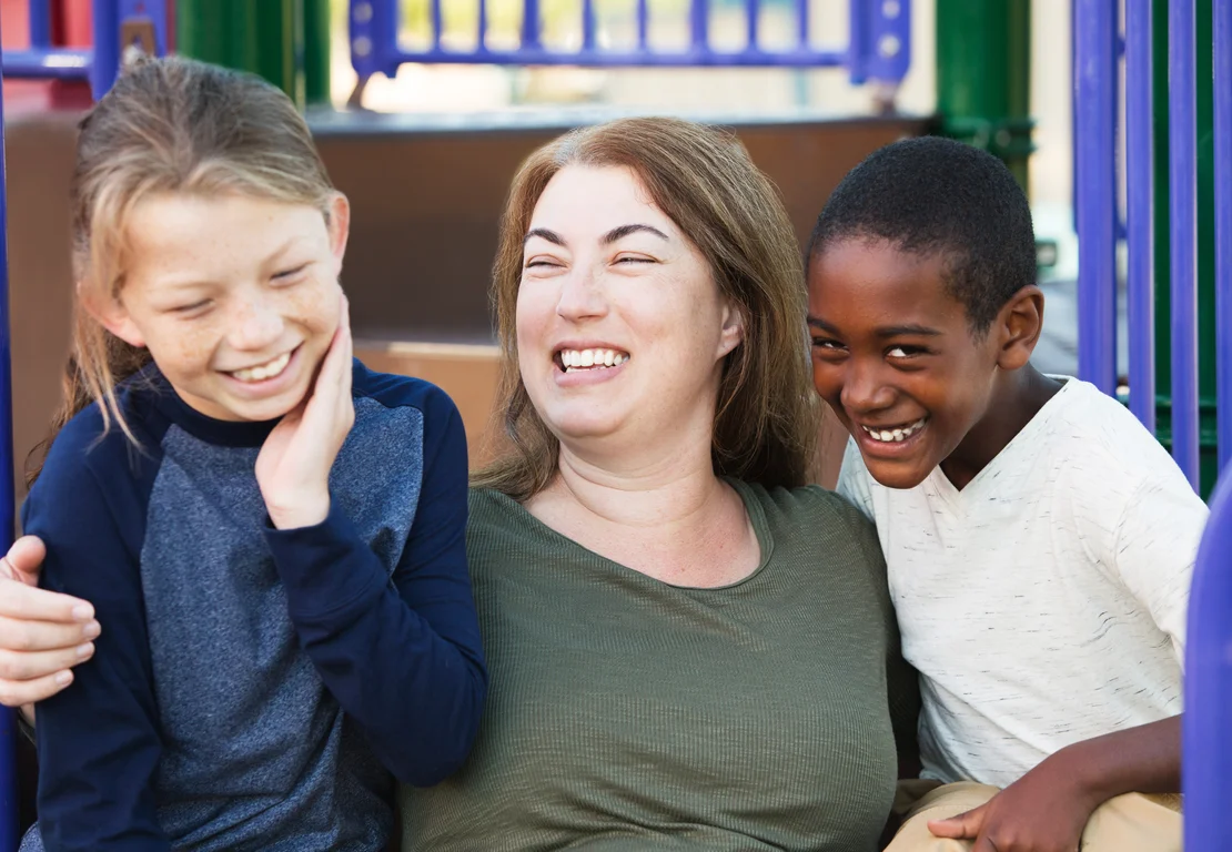 A mother laughing joyfully with her two children at a park playground